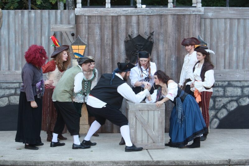 The twelfth night play stage with multiple cast members dressed up watching an actor and actress arm wrestle on a wooden box.